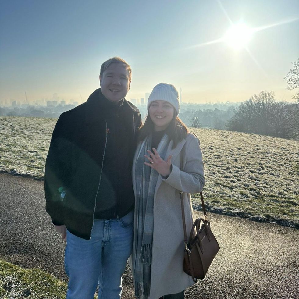 A man and a woman stand on the side of hill with the sun behind them. London can be seen in the background. They are both smiling. The woman has her hand raised, displaying a ring on her left hand.